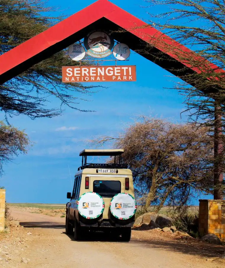 A Tanzania Safari Experience 4x4 Land Cruiser parked at the Serengeti National Park entrance gate under a bright blue sky.